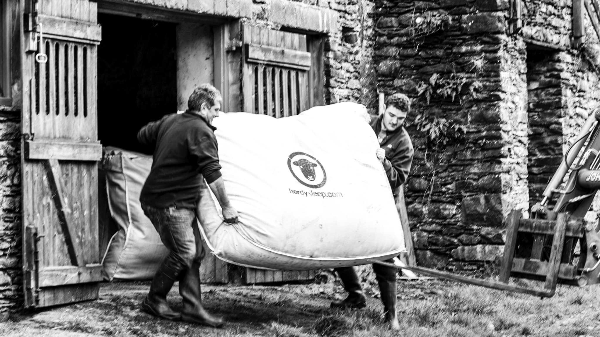 Two Lake District fell farmers carry a heavy woolsack filled with Herdwick wool into a barn, ready for collection by Herdysleep. Photo ©Spencer Hannah.